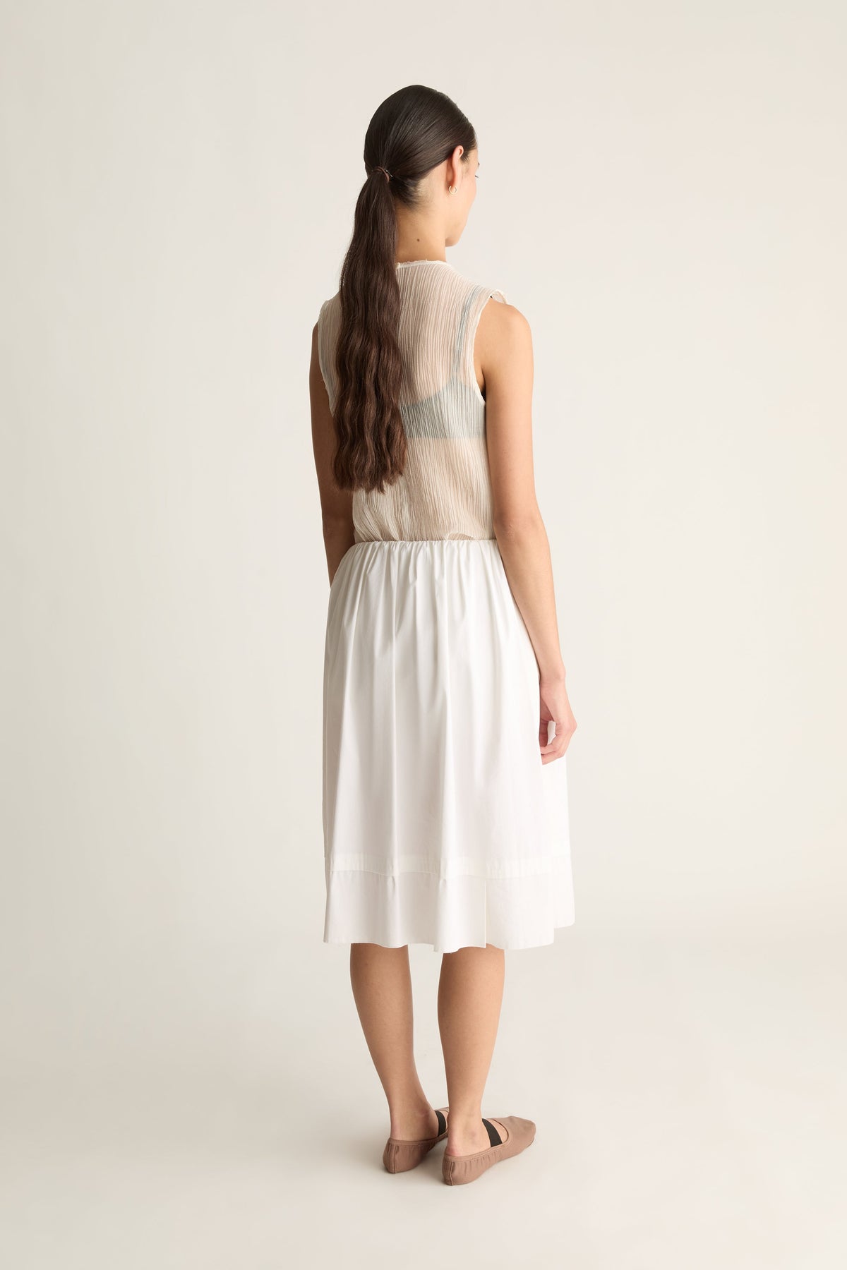 Back view of a woman with a ponytail in a white elastic-waist skirt with tiered hem and a sheer sleeveless top
