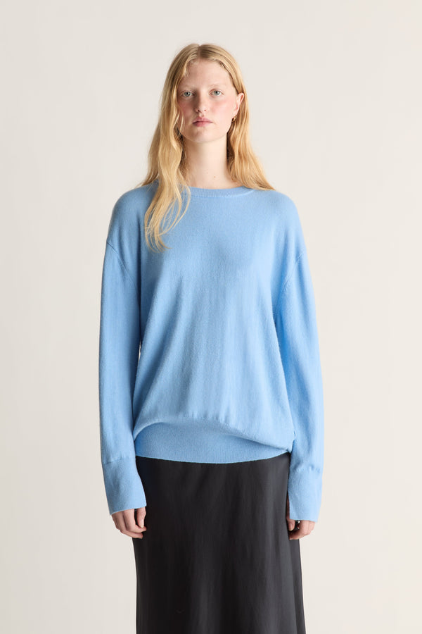 A blonde model wearing a light blue knit sweater and a black skirt, standing against a plain light beige studio background.