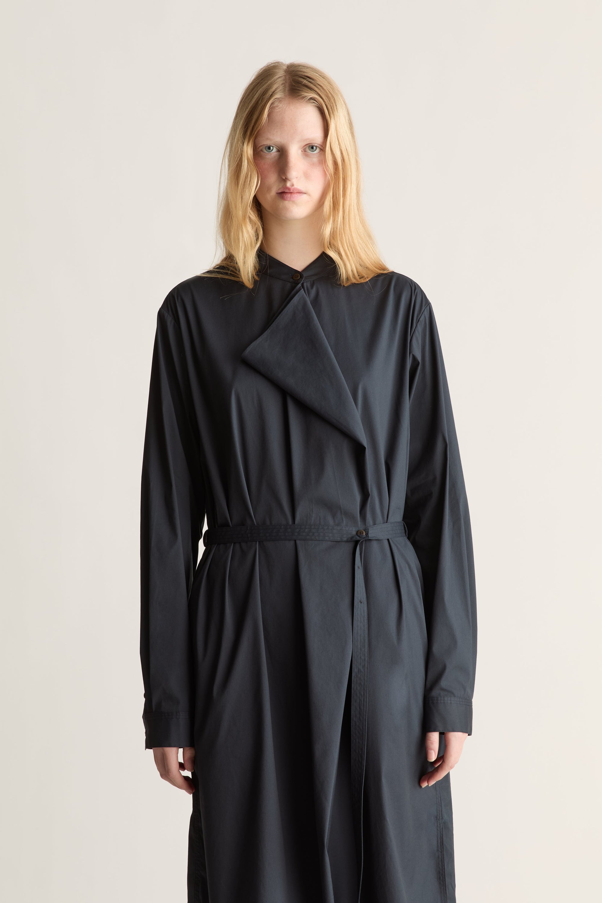 A model wearing a navy long-sleeve wrap-style dress with a tie waist, photographed against a plain light beige studio background.