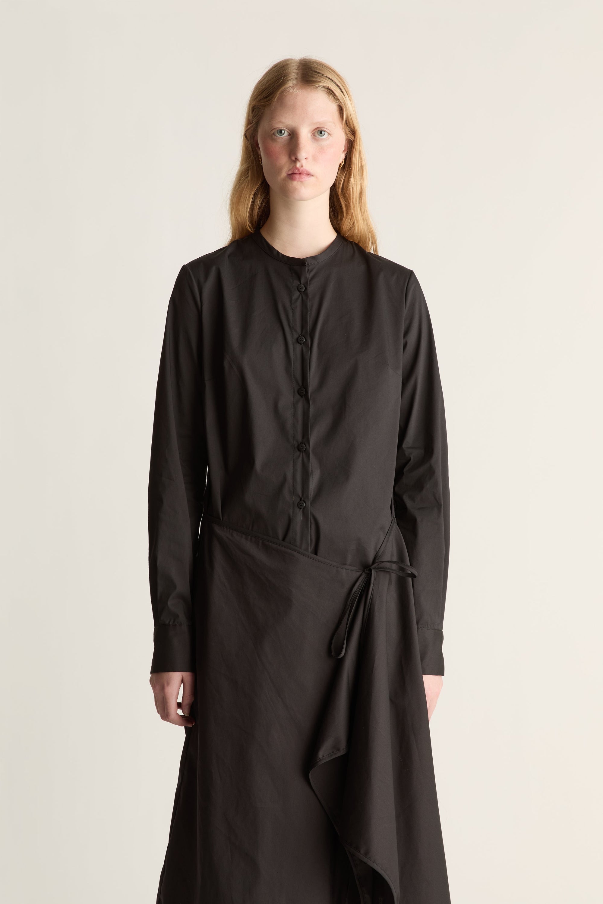 Close up of a woman wearing a black cotton long sleeve wrap dress against a white studio background. 