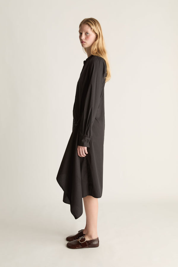 A woman modeling a loose, long black dress with a high-low asymmetrical hem and draped sleeves, paired with dark brown leather loafers, against a plain light beige studio background.