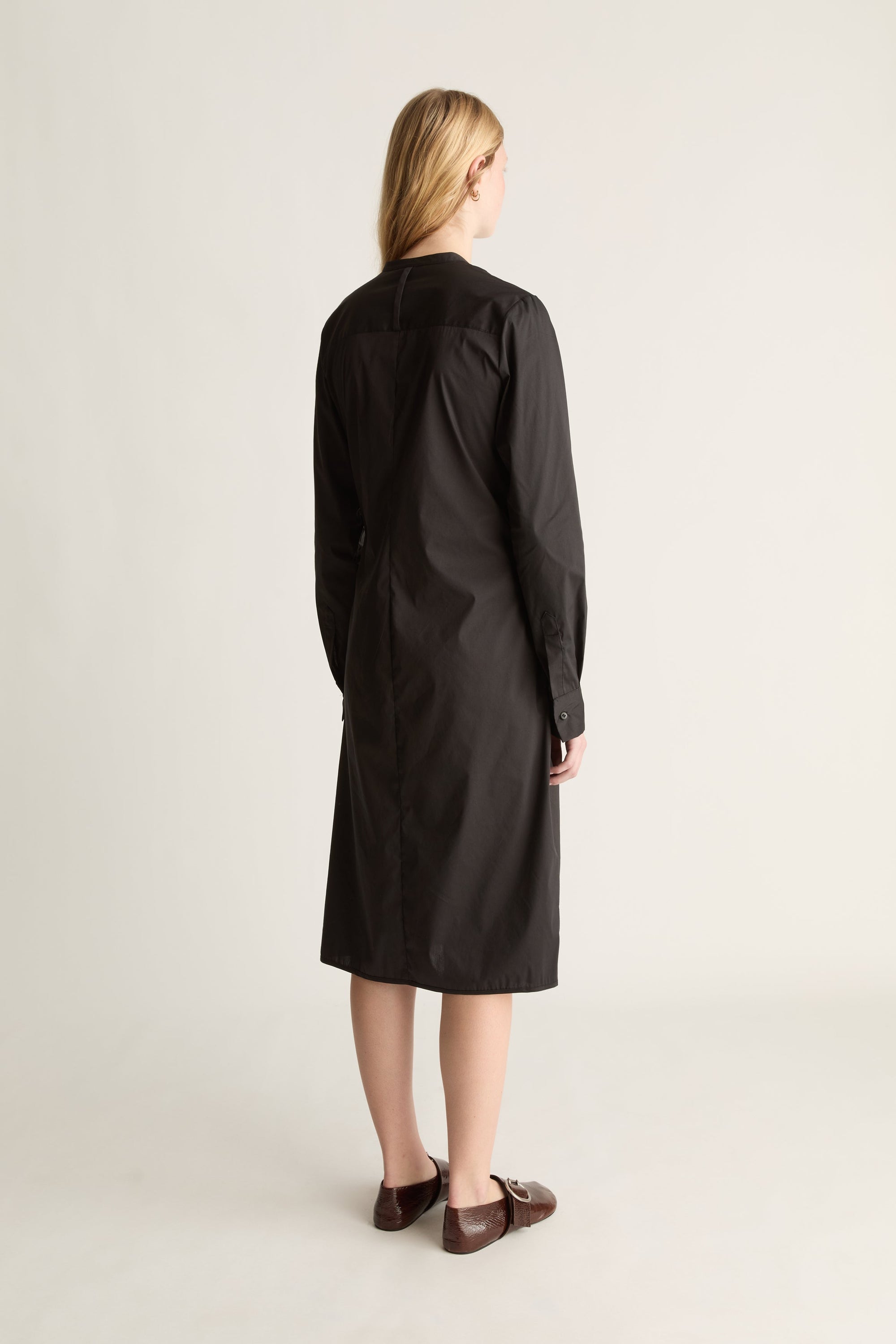 A woman stands with her back to the camera wearing a long black dress and brown leather loafers, photographed against a plain light beige studio background.