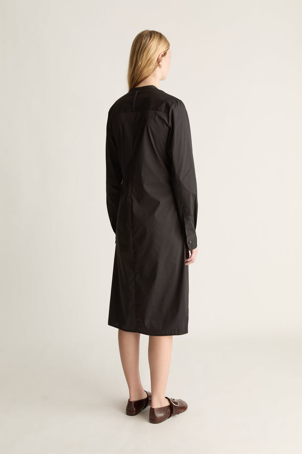 A woman stands with her back to the camera wearing a long black dress and brown leather loafers, photographed against a plain light beige studio background.