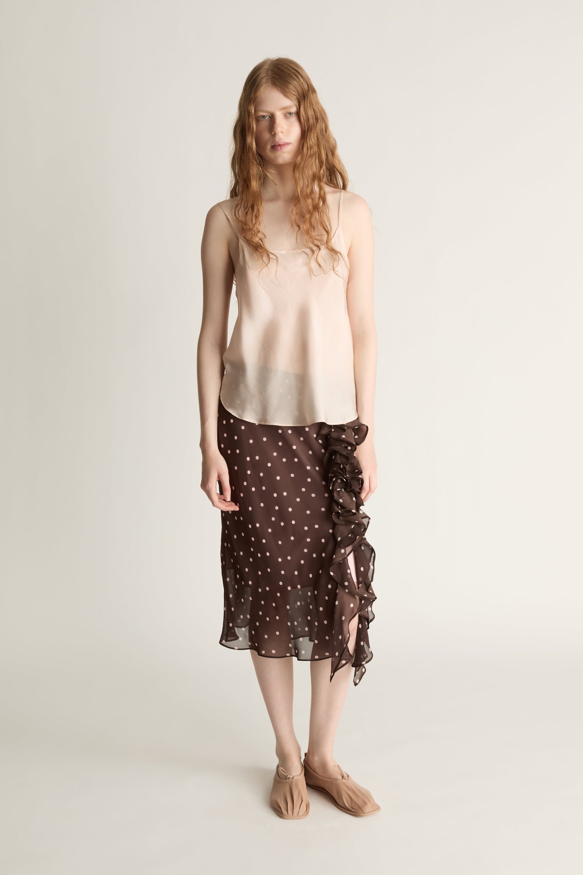 A woman wearing a beige cami top and brown and pink polka dot skirt on a plain background