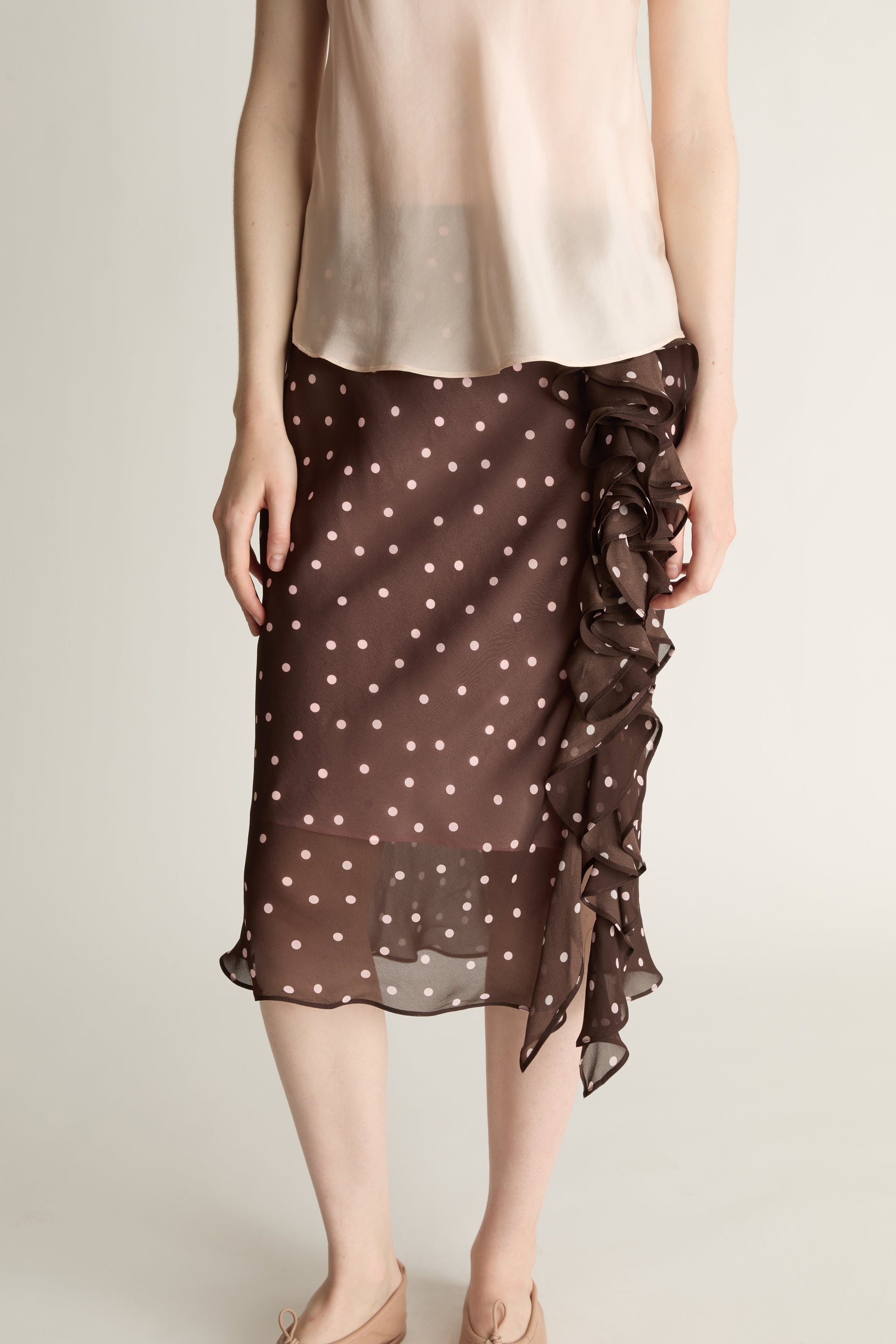 A woman wearing a beige cami top and brown and pink polka dot skirt on a plain background