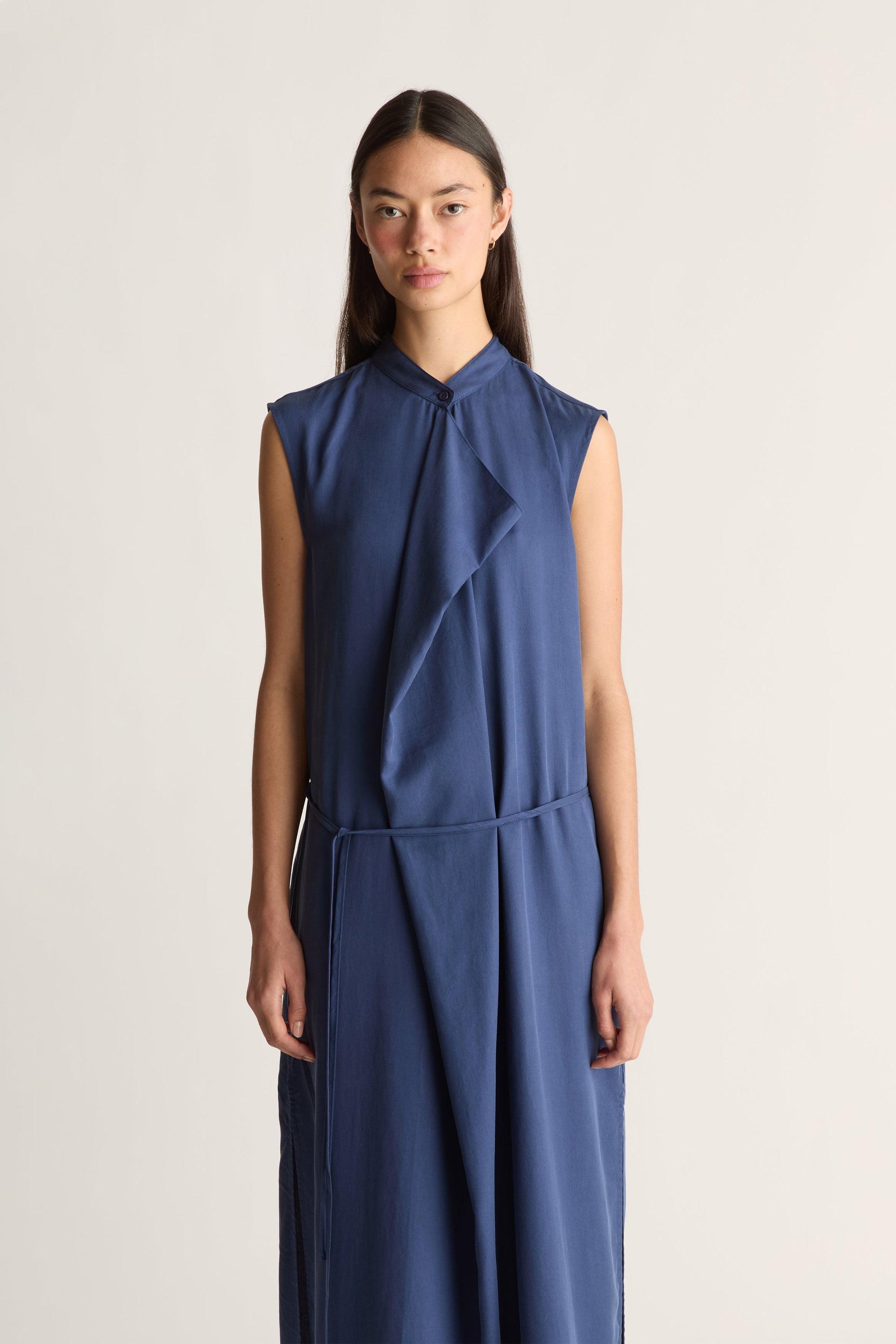 Close up of a woman wearing a sleeveless, high-neck blue dress with a draped wrap-style front and a matching tie belt, set against a plain light background.