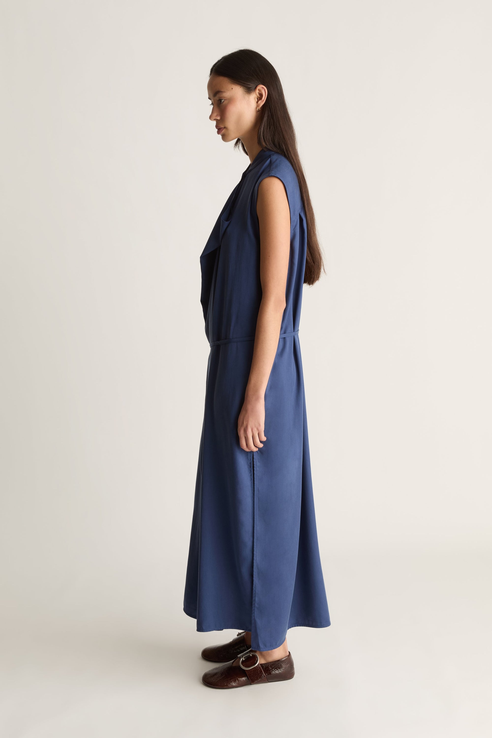 A woman stands in profile wearing a sleeveless navy blue midi dress with a draped neckline and brown leather loafers, photographed against a plain light beige studio background.