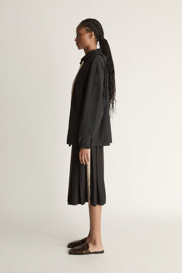 Woman wearing a black jacket with a white zipper and a black pleated skirt on a light background