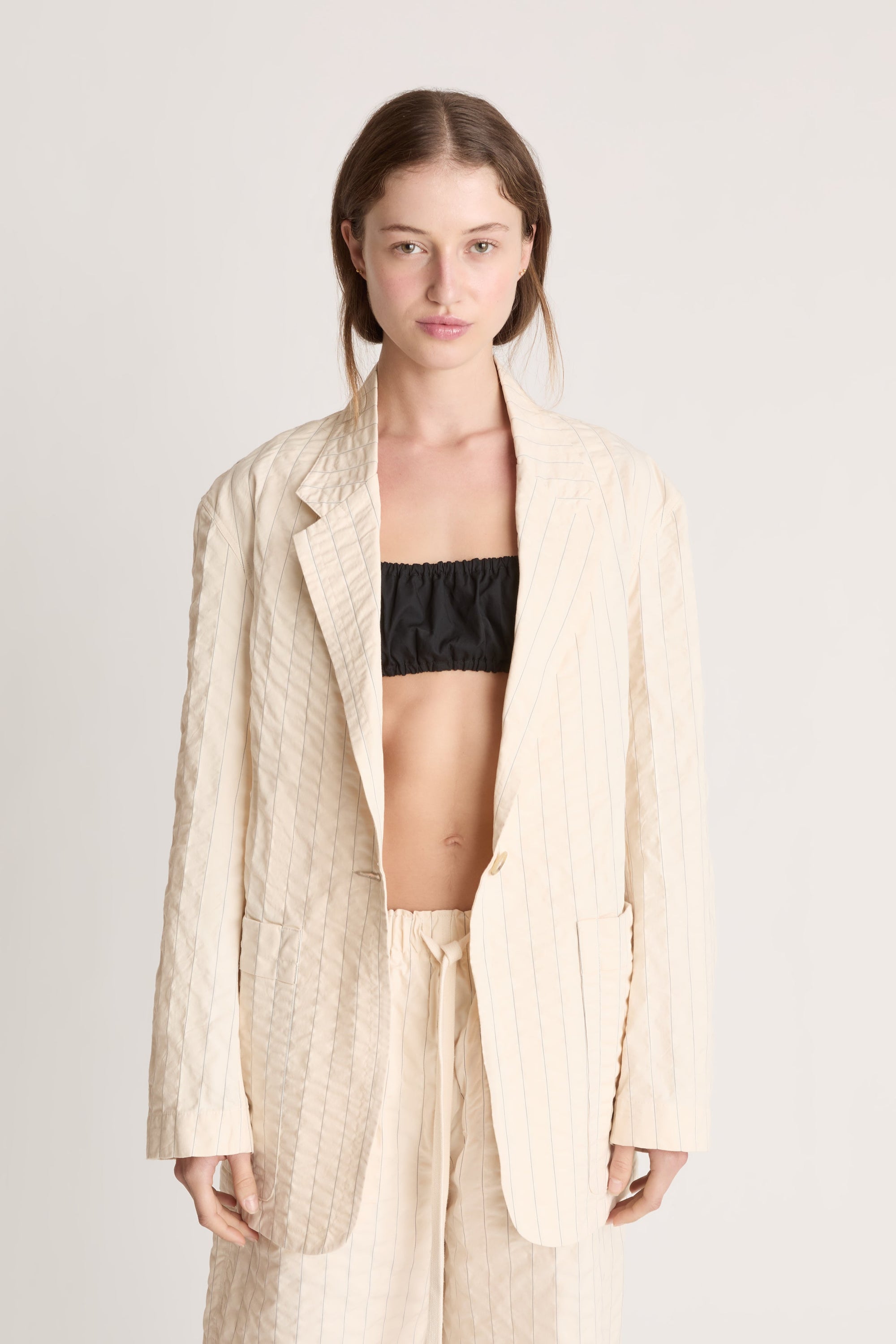 Woman wearing a cream blazer and shorts with a black bralette underneath on a white background.