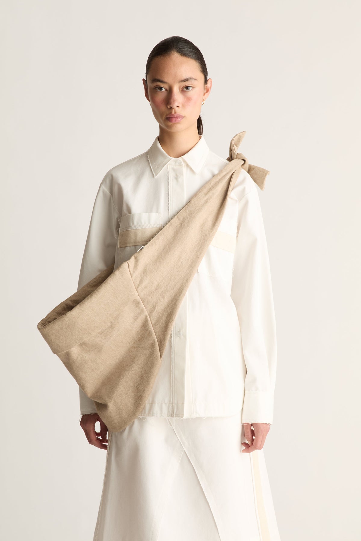 Front view of a woman wearing a white button-up shirt and white skirt with a beige knotted cross body bag against a white studio background
