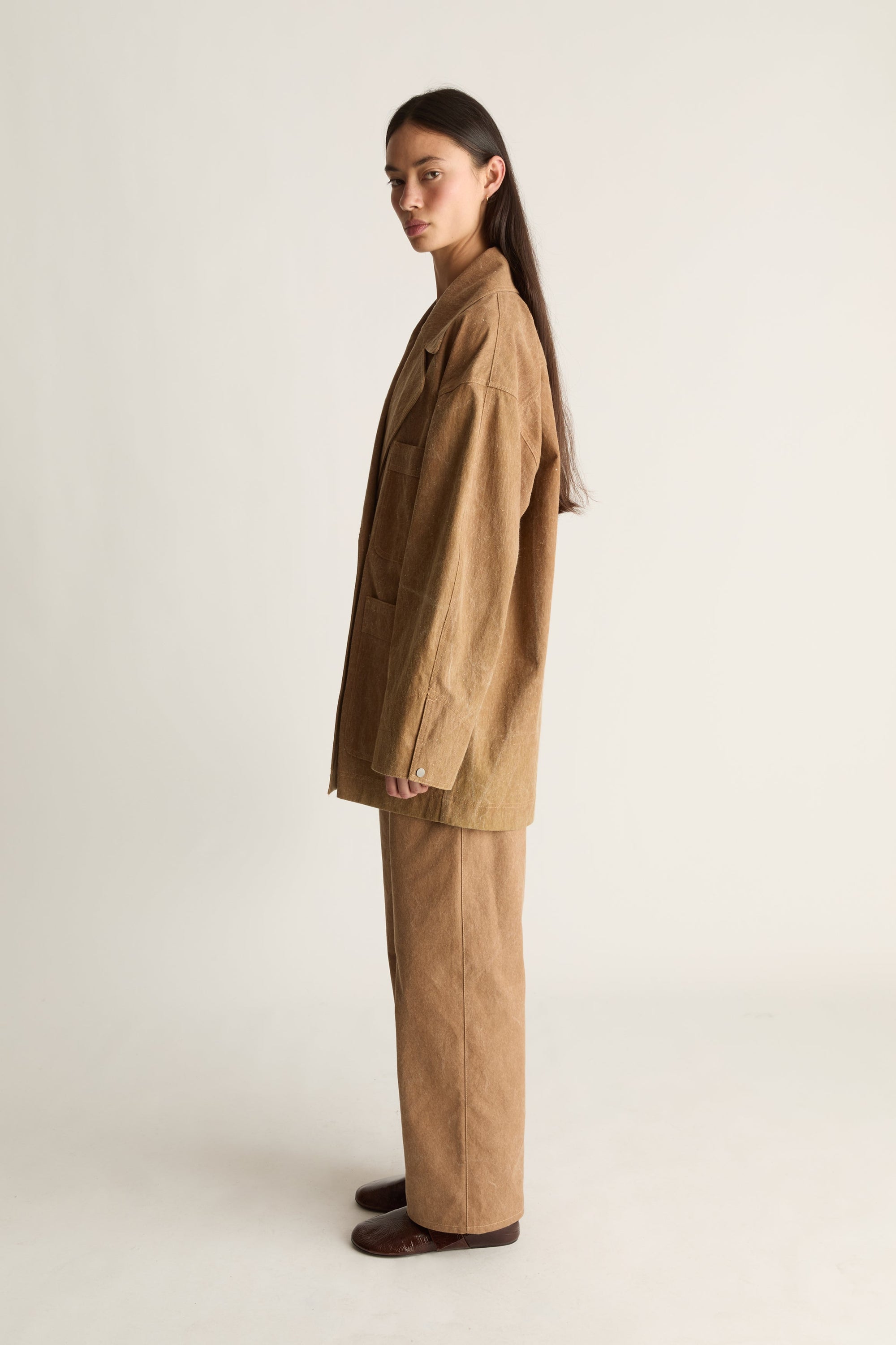 Woman wearing a brown coat and pants on a white background, facing the side but looking toward the front.