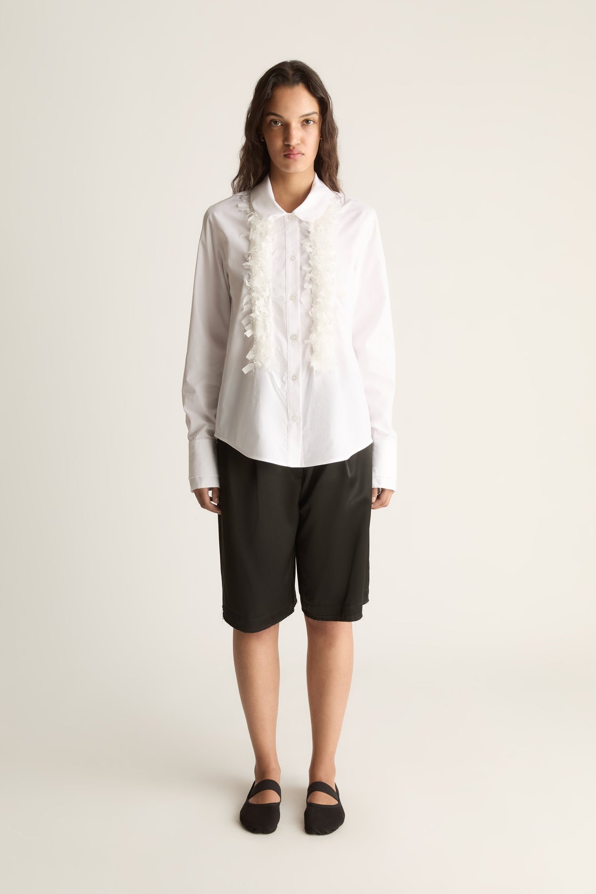Front view of a woman wearing a white long-sleeve shirt with ruffle trim and black knee-length shorts against a white studio background