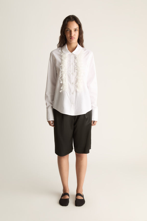 Front view of a woman wearing a white long-sleeve shirt with ruffle trim and black knee-length shorts against a white studio background