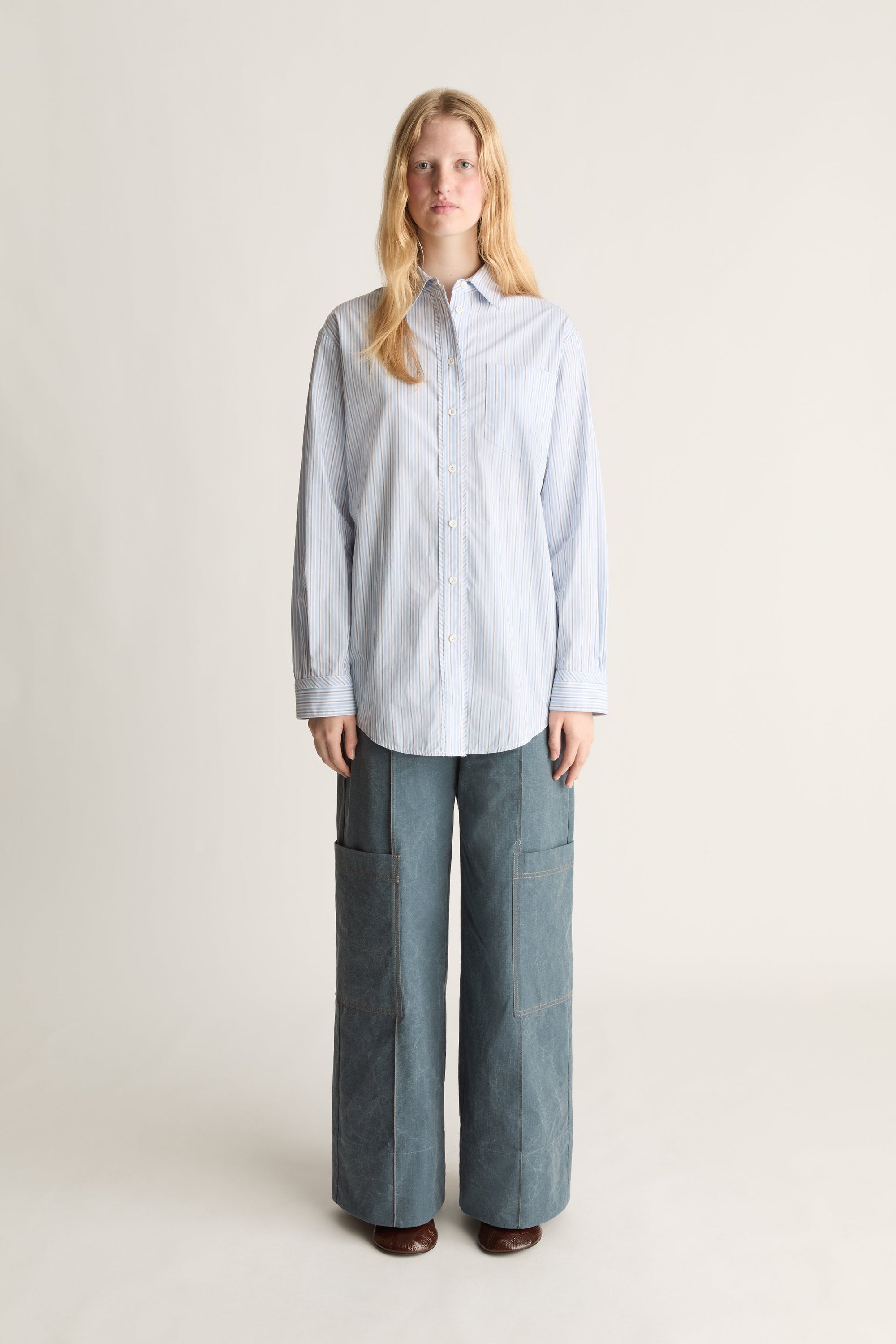 A woman stands against a plain light beige studio background wearing a light blue and white vertically striped button-up shirt and wide-leg blue pants.