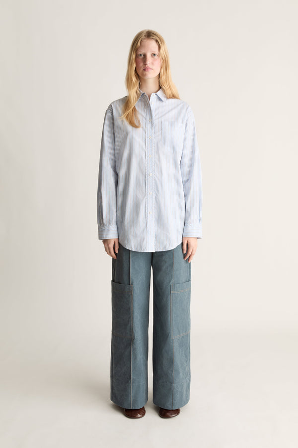 A woman stands against a plain light beige studio background wearing a light blue and white vertically striped button-up shirt and wide-leg blue pants.