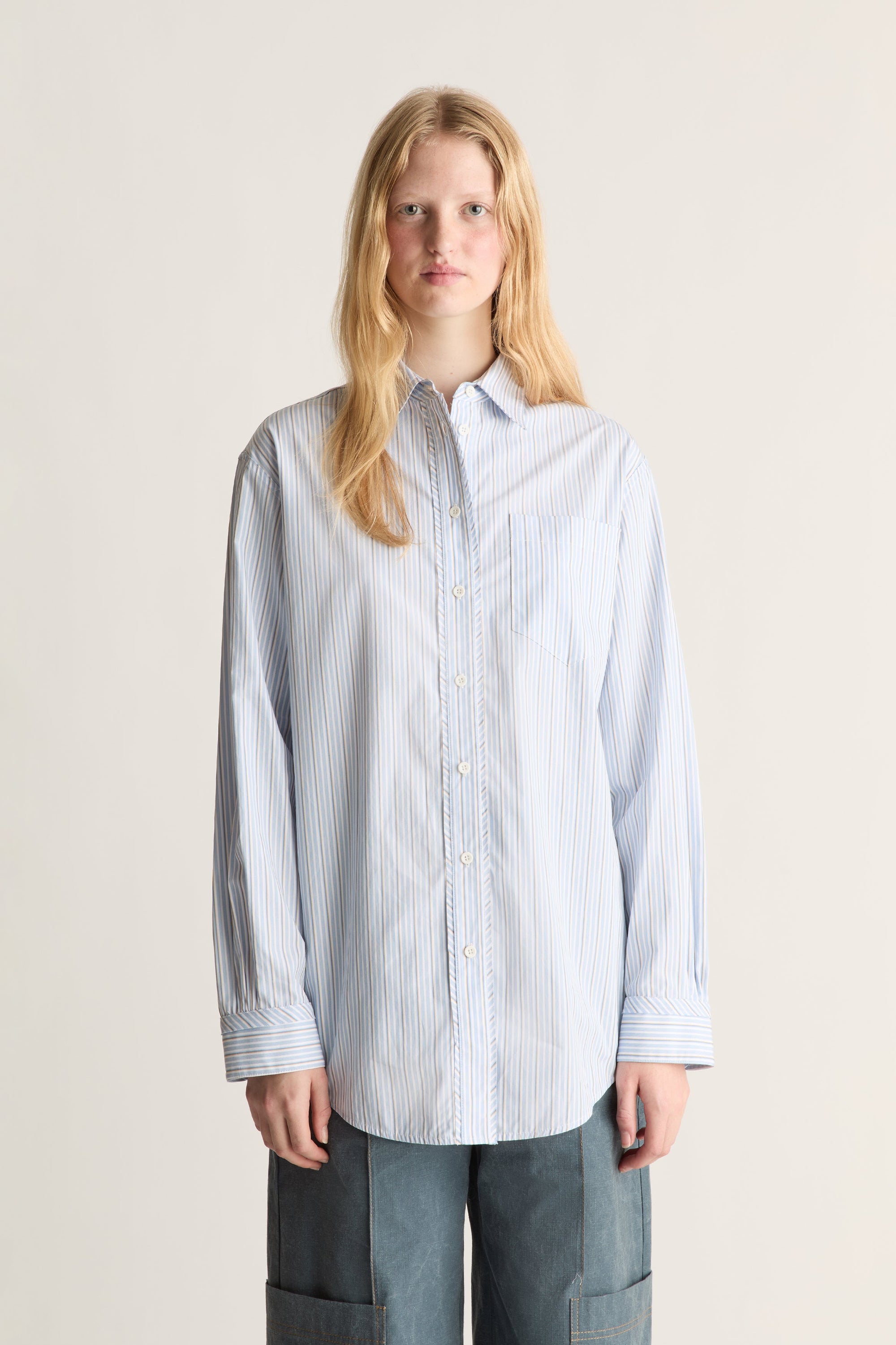 Close up of a  Woman wearing a light blue and white vertically striped button-down shirt with dark blue pants, standing against a plain light beige background.