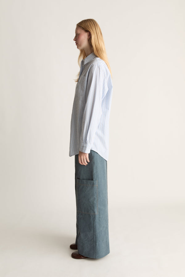 A woman stands in profile wearing a light blue long-sleeve button-up shirt and wide-leg blue jeans against a plain off-white studio background.
