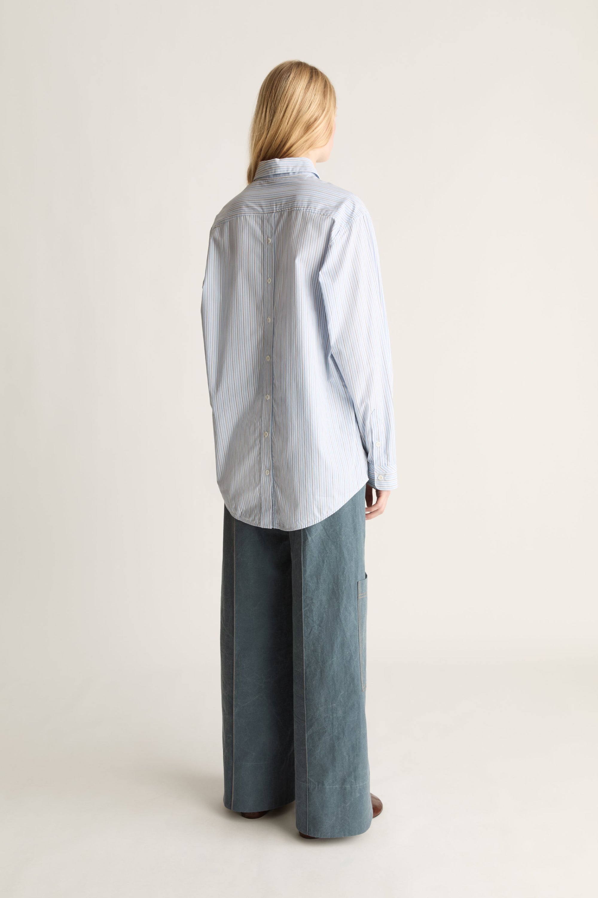 A woman shown from the back wearing a light blue and white striped button-up shirt and wide-leg slate blue pants, standing against a plain off-white studio background.