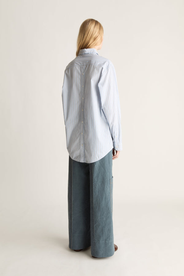 A woman shown from the back wearing a light blue and white striped button-up shirt and wide-leg slate blue pants, standing against a plain off-white studio background.
