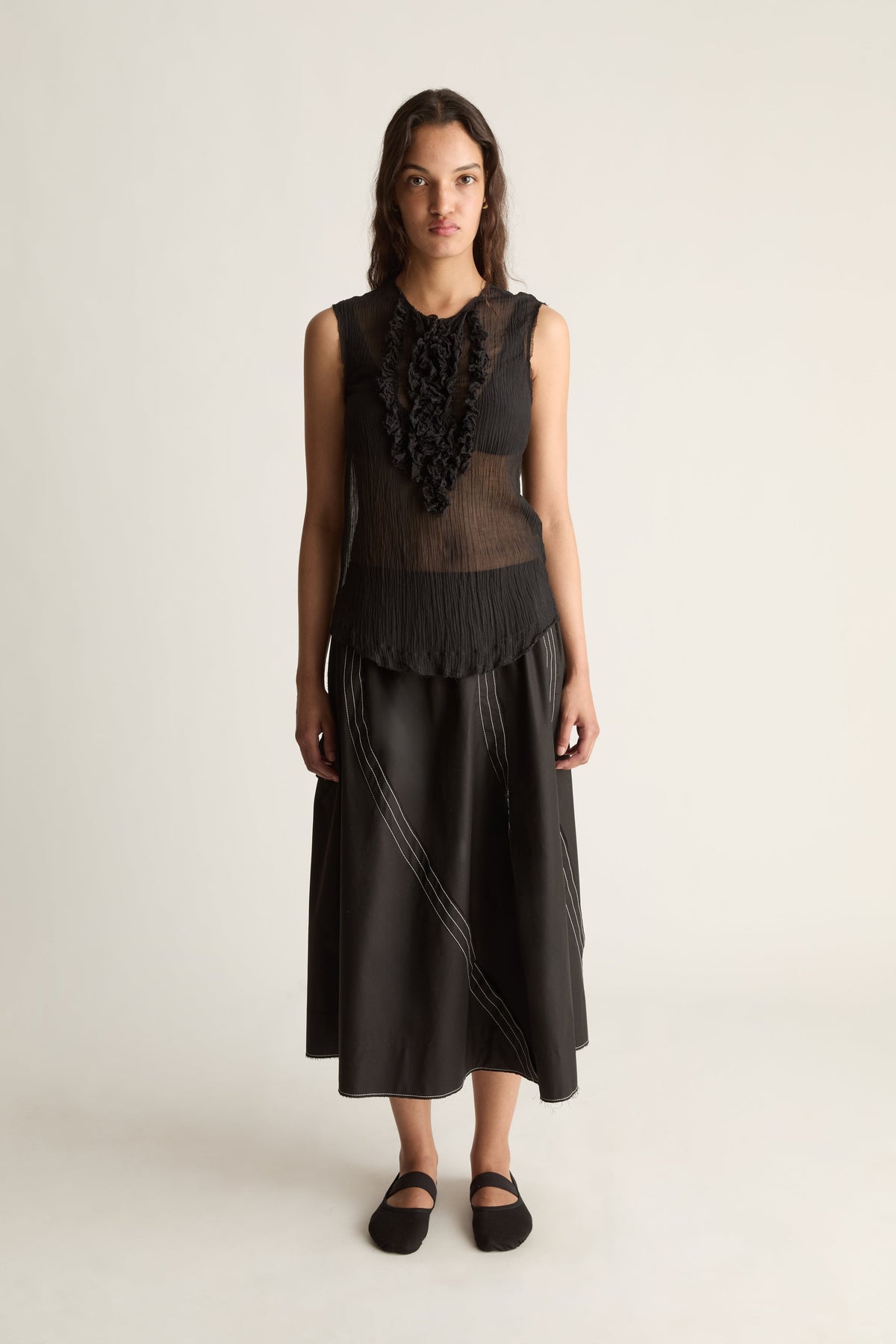 Front view of a woman wearing a sheer black sleeveless ruffle top and black midi skirt with white stitching against a white studio background