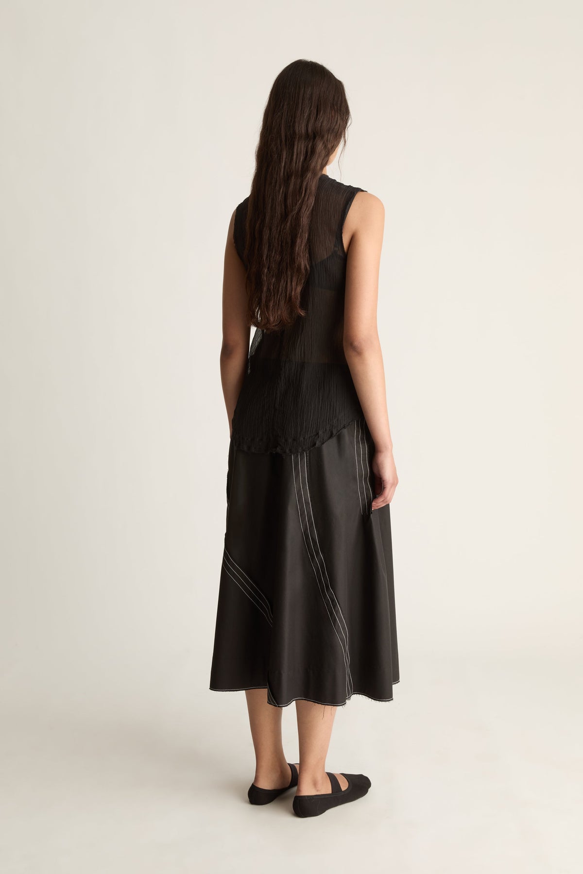Back view of a woman with long hair in a sheer black pleated top and black skirt with white topstitching