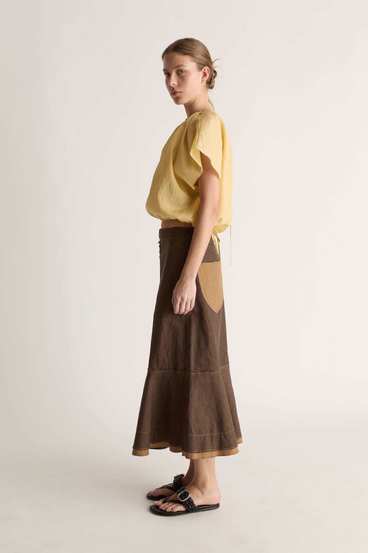 Side profile of a woman in a yellow short-sleeve gathered top and brown midi skirt with tan side pocket
