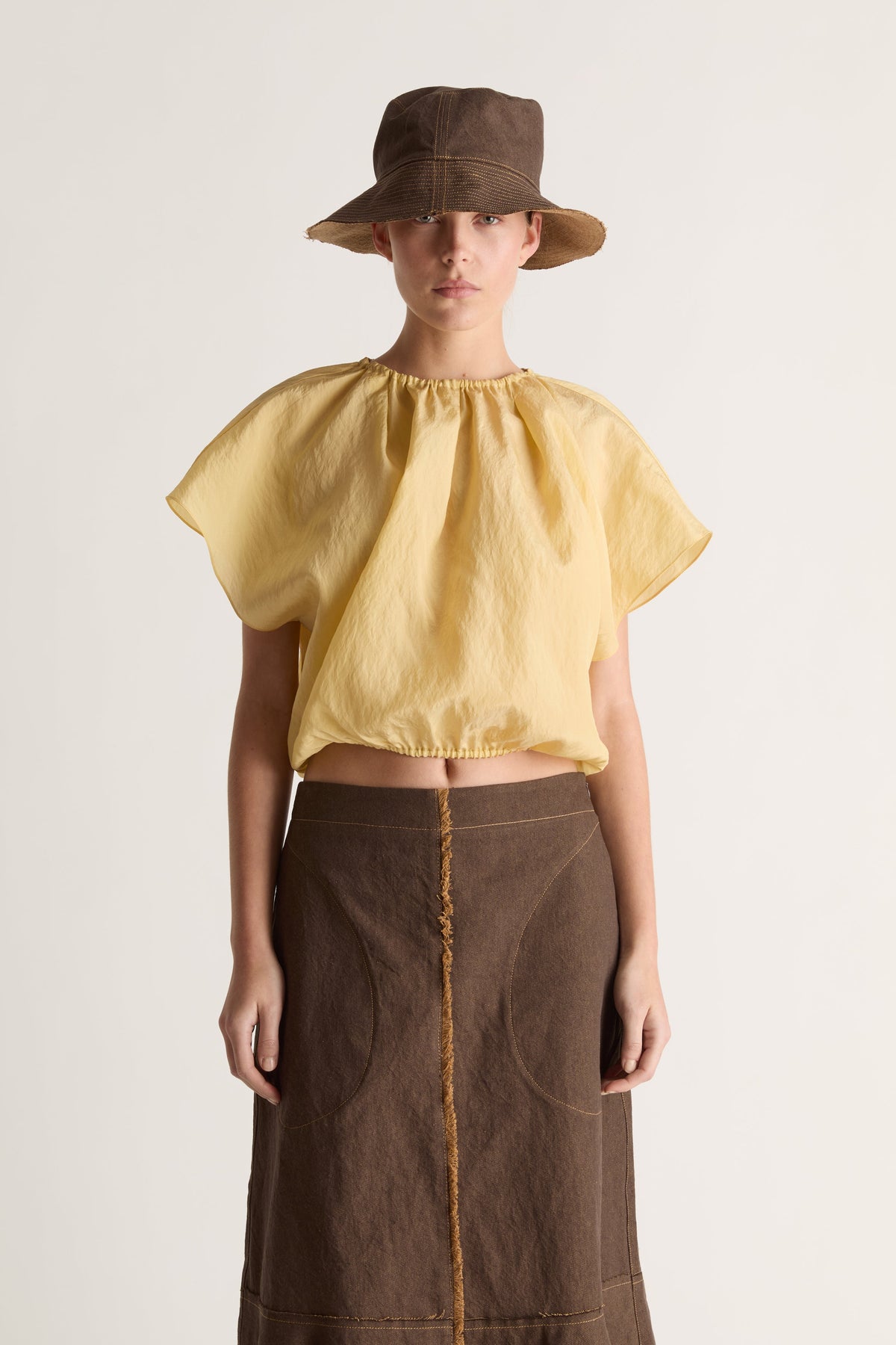 Close-up of a woman in a brown bucket hat, yellow gathered cropped top and brown skirt with contrast stitching