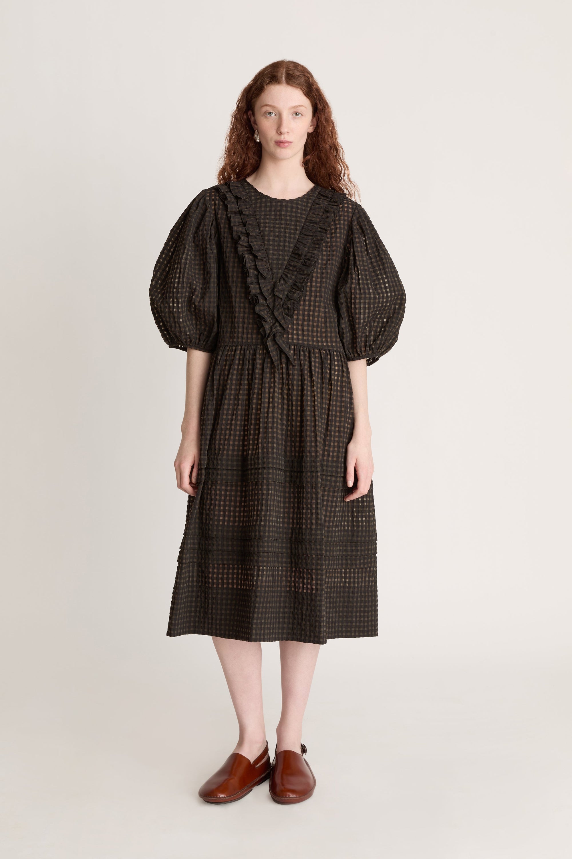Woman wearing a dark checkered dress  with puffy sleeves with ruffled details on a white background