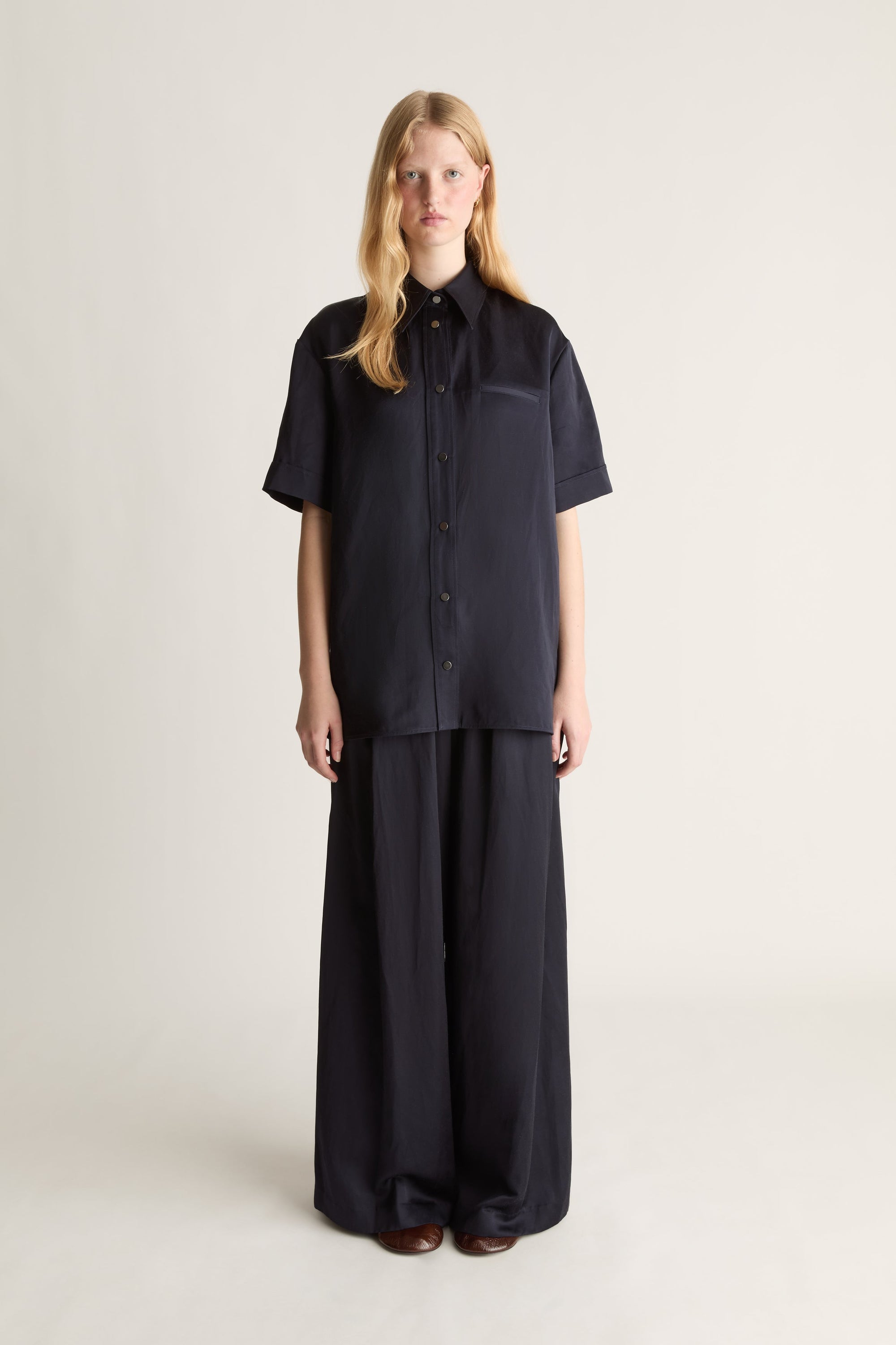 A woman stands against a plain light beige studio background wearing a dark navy short-sleeve button-front shirt with a chest pocket and matching dark wide-leg pants.