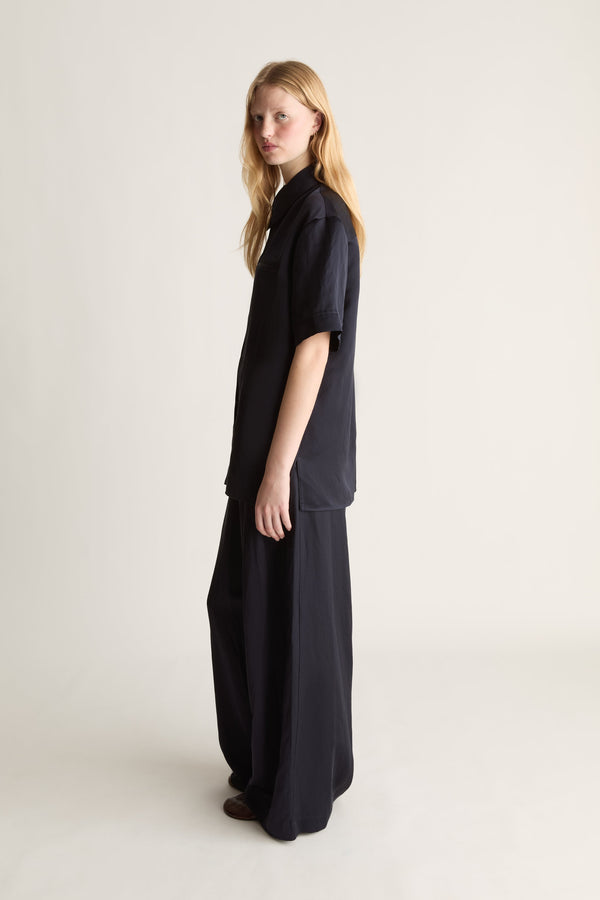 Side view of a woman modeling a navy short-sleeve collared shirt and matching wide-leg trousers against a plain light beige studio background.