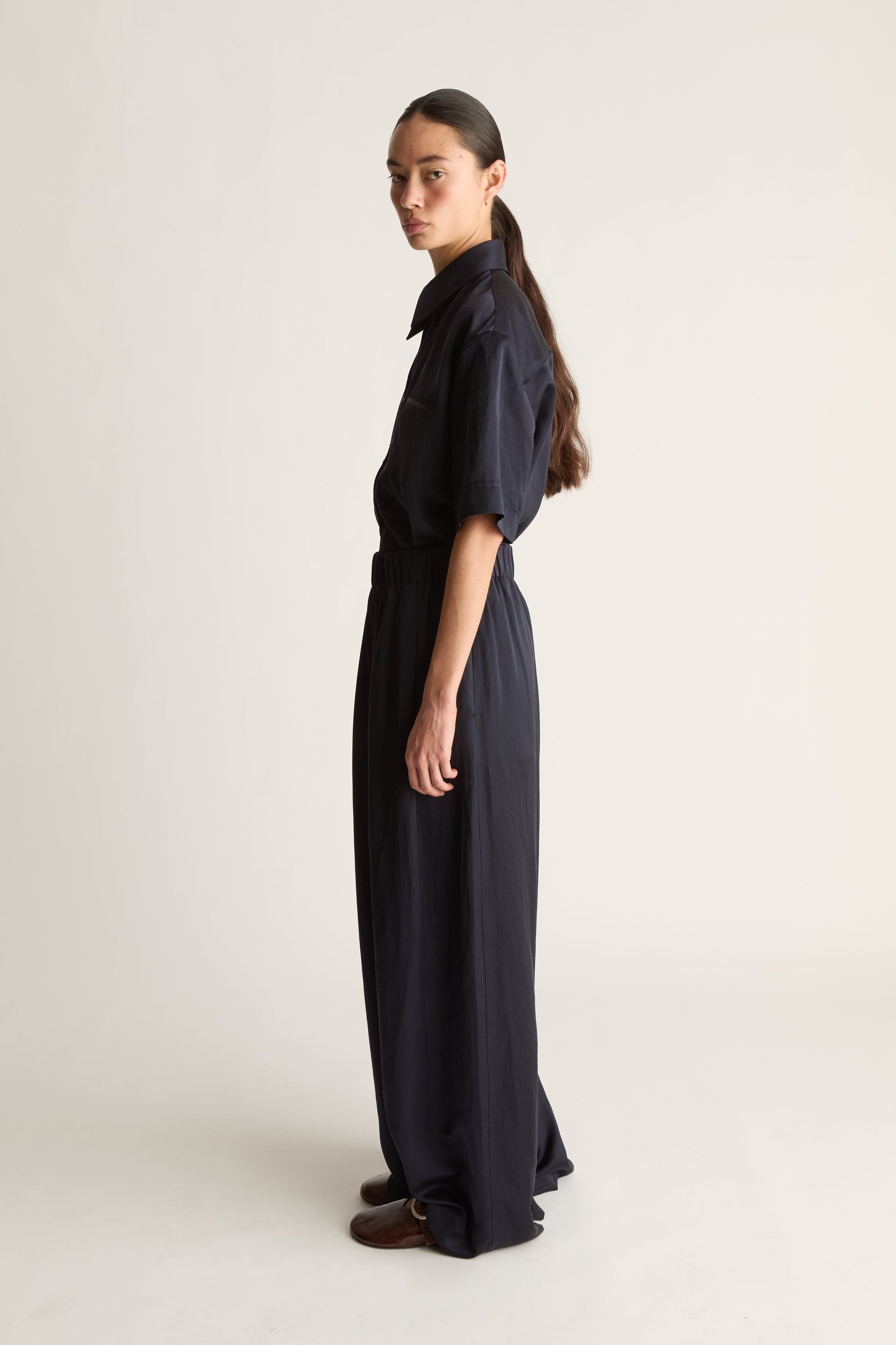 A model stands in profile wearing a dark navy blue long-sleeve shirt and matching wide-leg trousers, photographed against a plain light beige studio background.