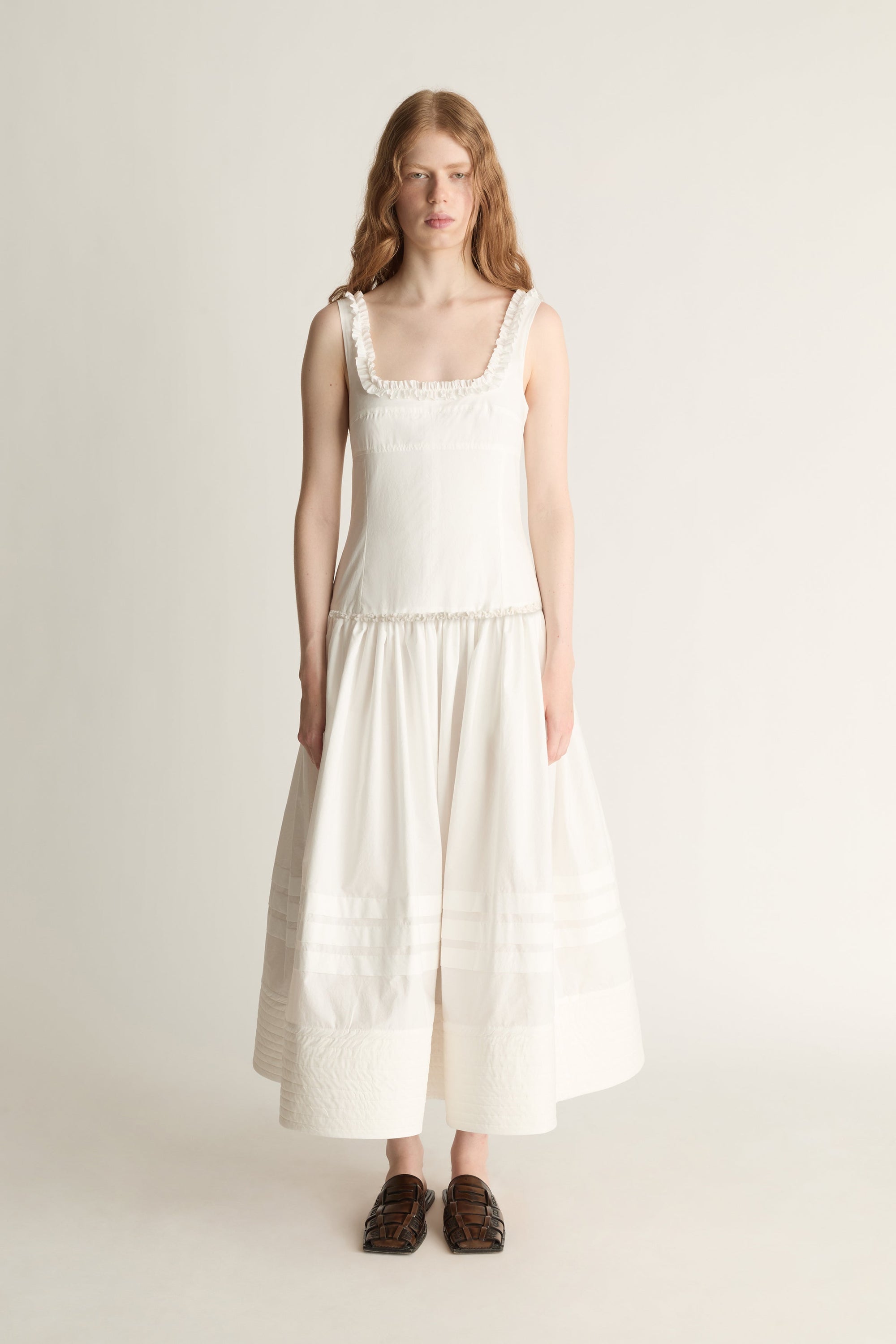 A woman stands against a plain light beige studio background wearing a sleeveless cream dress with a square neckline, delicate lace trim at the shoulders and waist, and a tiered skirt. She is wearing dark brown loafers.