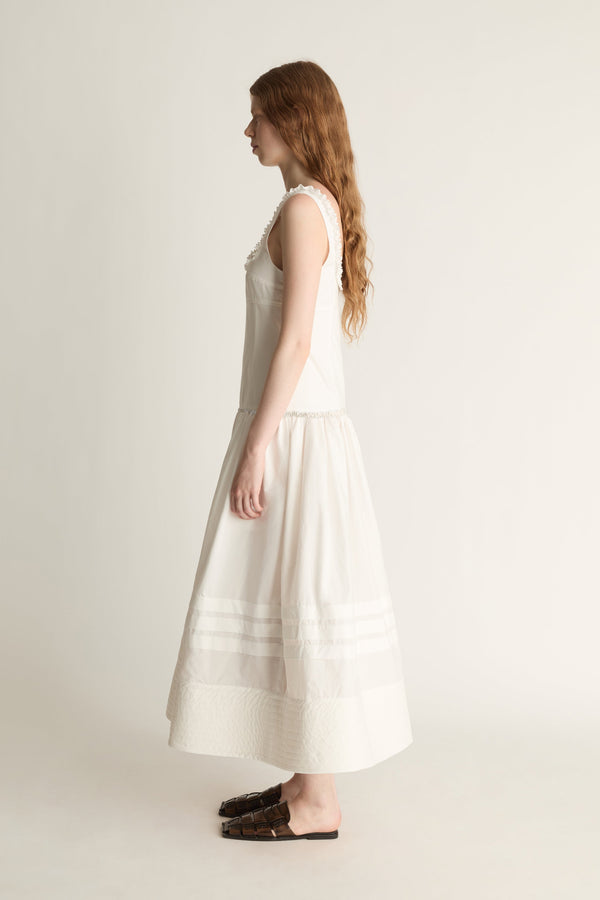 A woman in profile wearing a sleeveless cream-colored midi dress with horizontal embroidered stripes near the hem, paired with dark brown flat shoes, set against a plain light beige studio background.