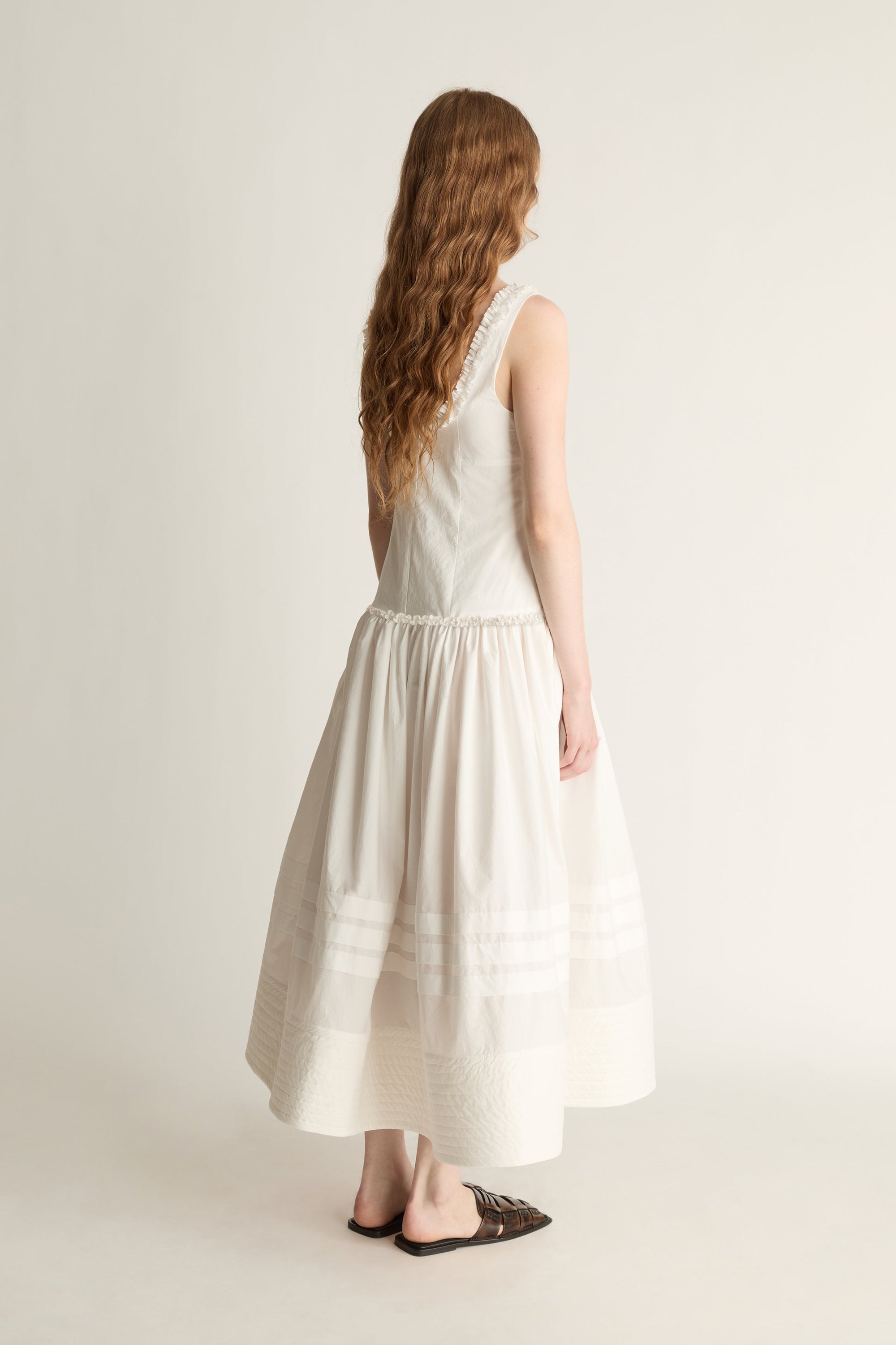 A woman shown from the back wearing a sleeveless, off-white midi dress with a flared skirt, paired with dark brown flat sandals, against a plain light beige studio background.