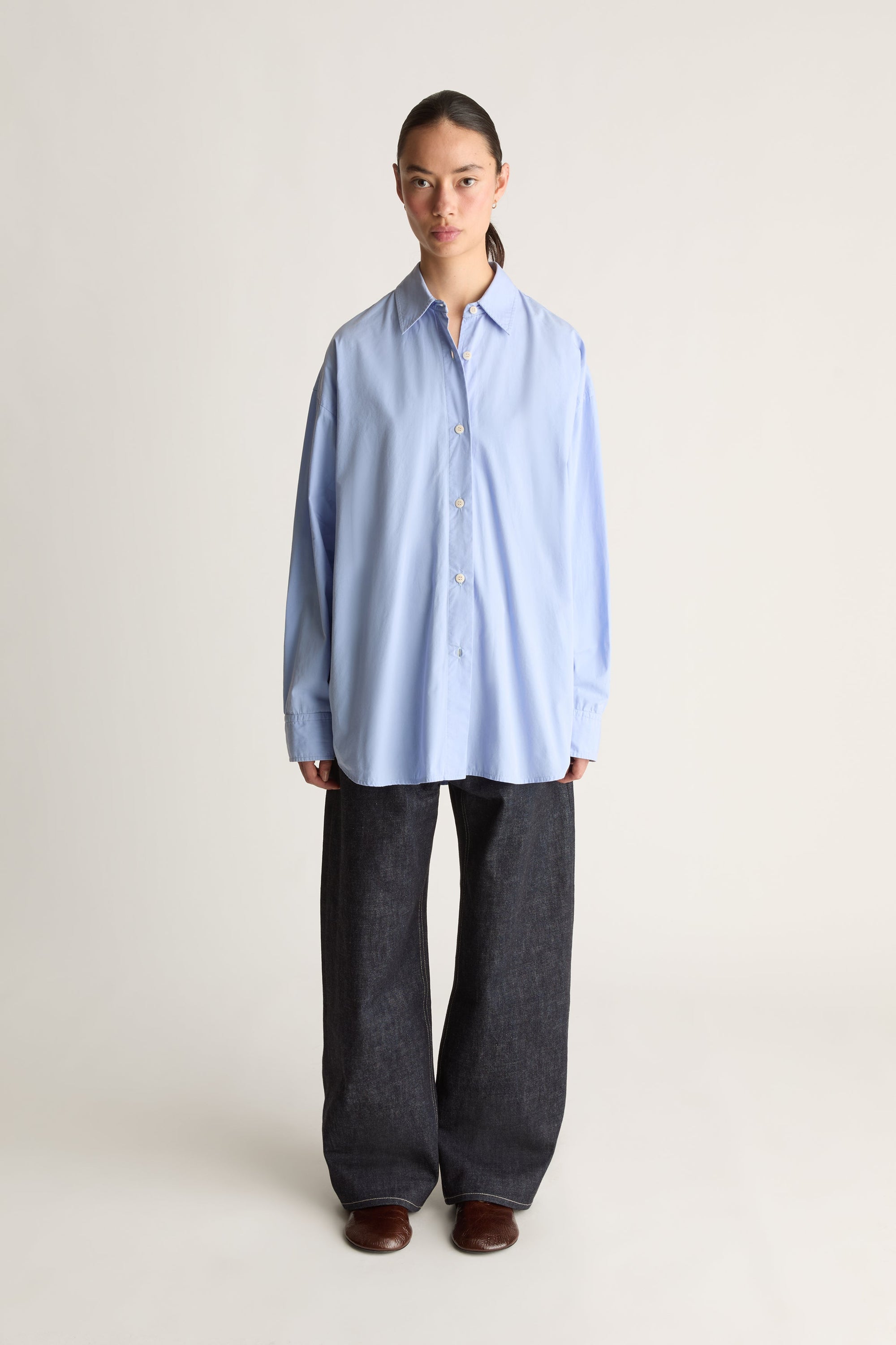 A model stands against a plain light beige studio background wearing an oversized light blue button-up shirt and very wide-leg dark charcoal gray trousers.
