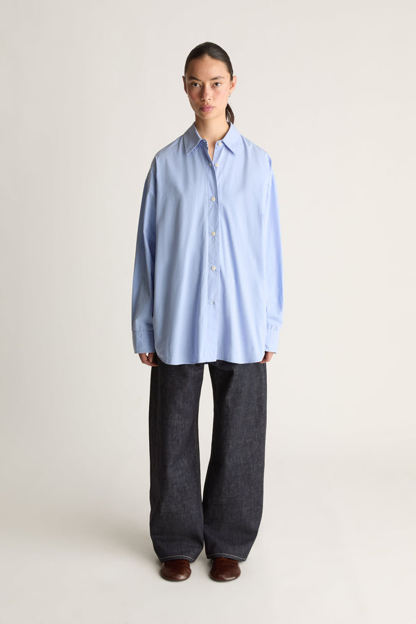 A model stands against a plain light beige studio background wearing an oversized light blue button-up shirt and very wide-leg dark charcoal gray trousers.