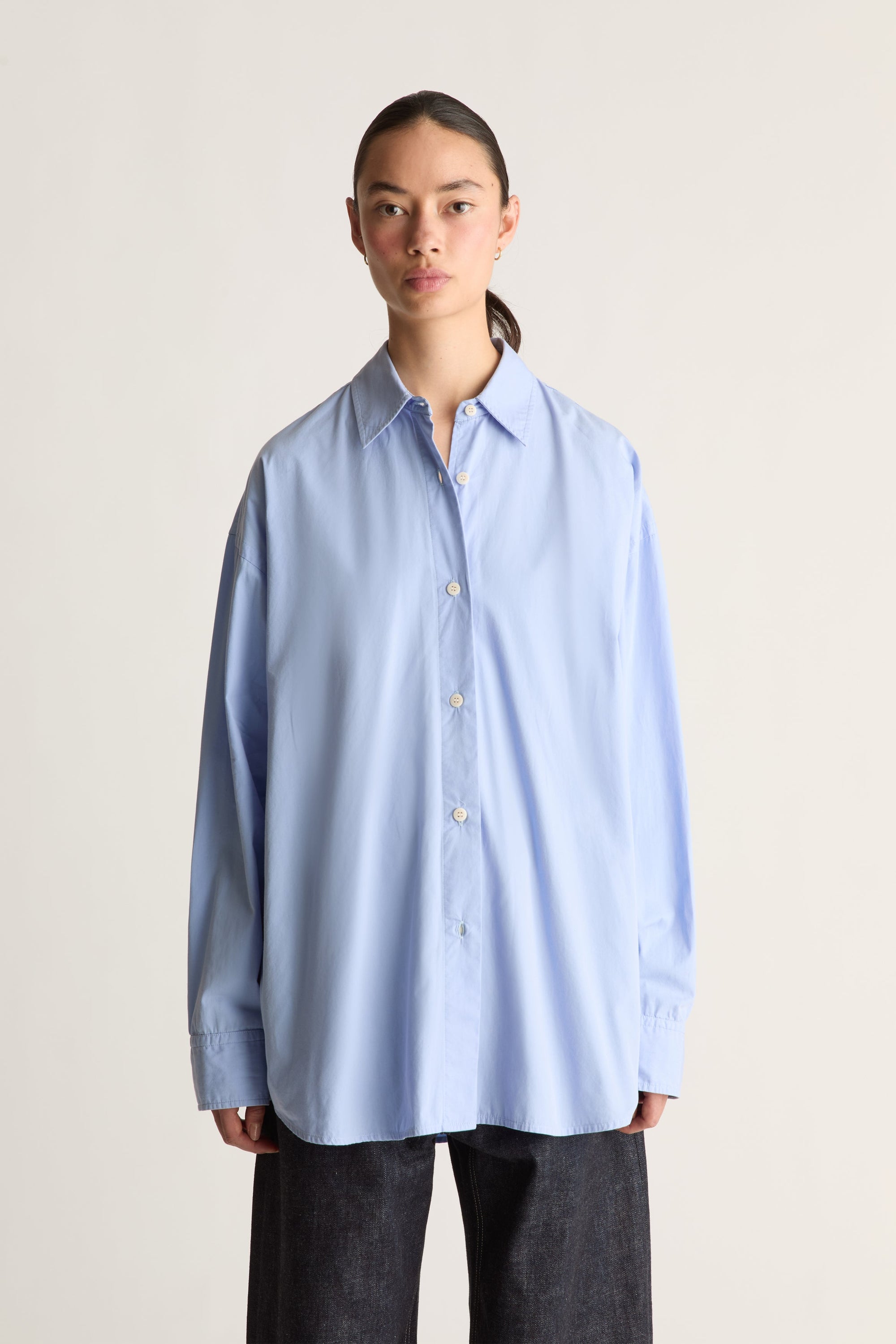 A model wearing an oversized light blue long-sleeve button-up shirt and dark charcoal pants, standing against a plain light beige studio background.
