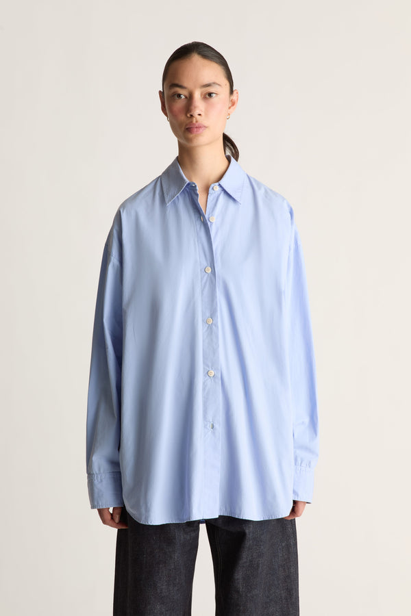 A model wearing an oversized light blue long-sleeve button-up shirt and dark charcoal pants, standing against a plain light beige studio background.