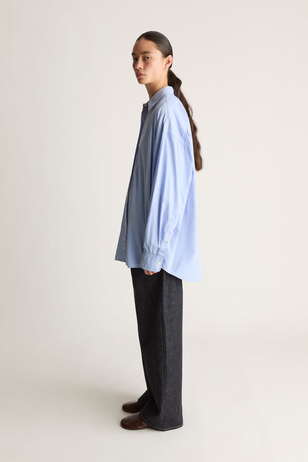 A model stands in profile wearing an oversized light blue long-sleeve button-up shirt, dark wide-leg jeans, and brown shoes against a plain light gray studio background.