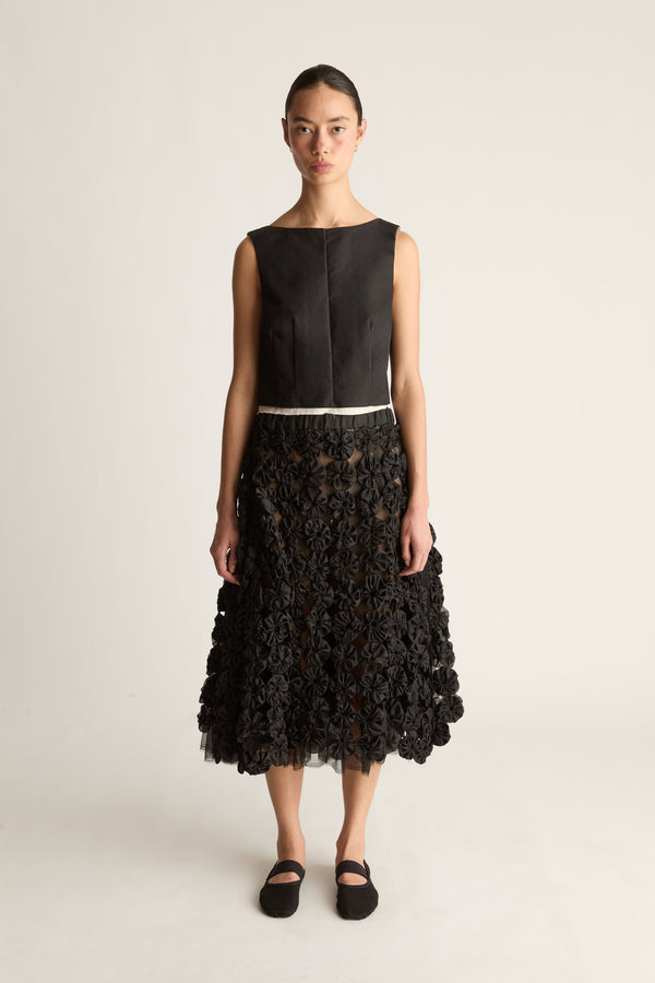 Front view of a woman wearing a black sleeveless top and black rosette midi skirt with tulle hem against a white studio background